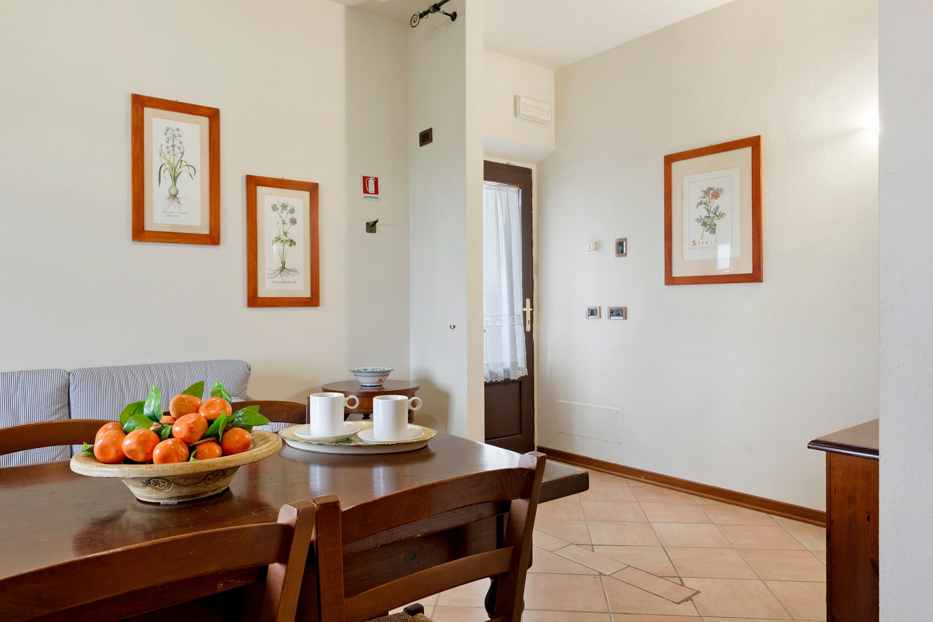 Un comedor con una mesa de madera dispuesta para dos personas, un cuenco de naranjas e ilustraciones botánicas enmarcadas en las paredes.