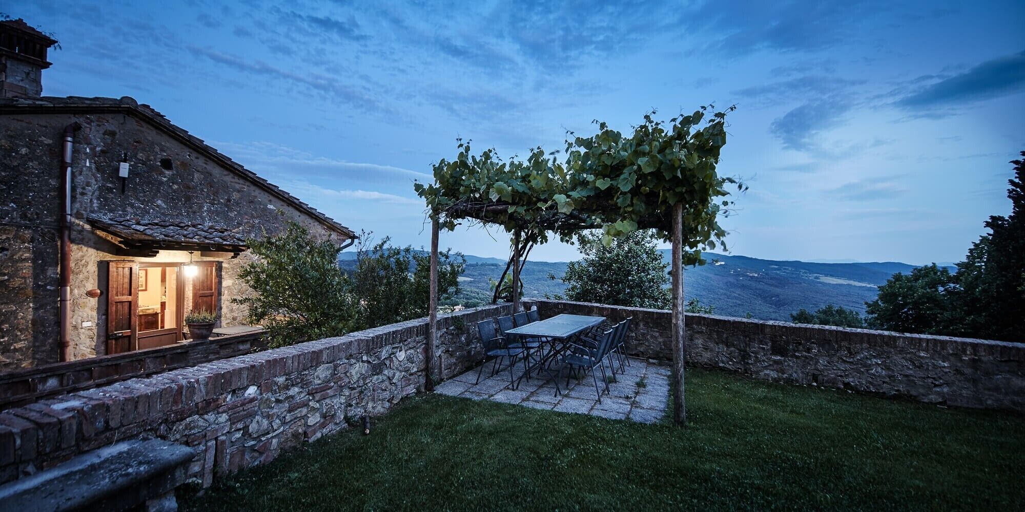Ein Steinhäuschen mit einer Terrassenfläche, komplett mit Tisch und Stühlen, einer mit Weinreben bewachsenen Pergola und Blick auf die Landschaft.