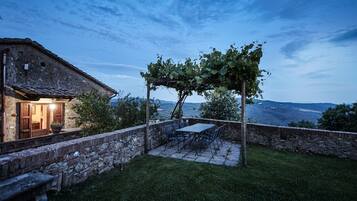 Ein Steinhäuschen mit einer Terrassenfläche, komplett mit Tisch und Stühlen, einer mit Weinreben bewachsenen Pergola und Blick auf die Landschaft.