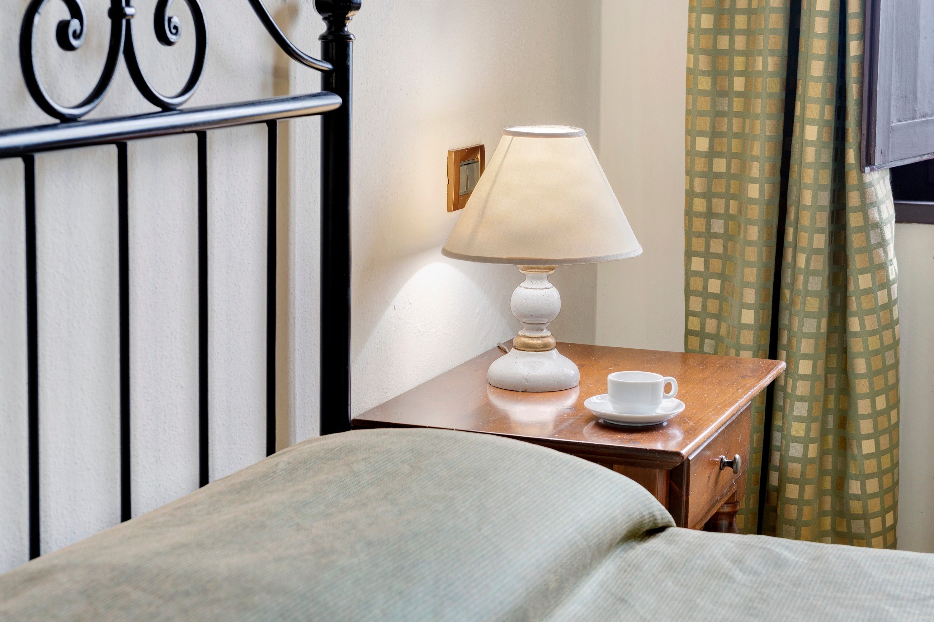 Ein Hotelzimmer mit einem Bett, einem Nachttisch mit Lampe, einer Tasse und einem Vorhang.