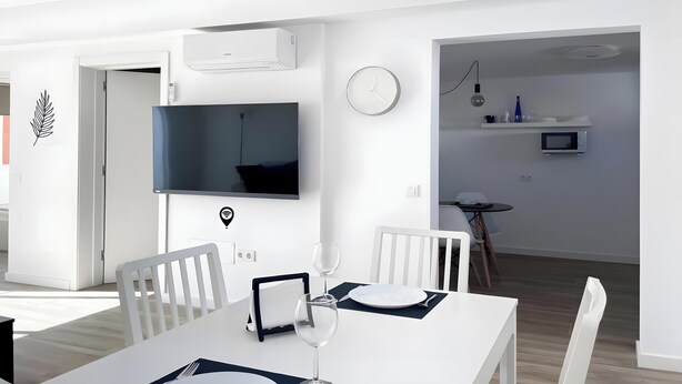A modern dining area with a white table, chairs, and a wine glass. A flat-screen TV is mounted on the wall above the table. - B-Rent La Azotea Zen – Your Relaxing Oasis in Corralejo (Corralejo)