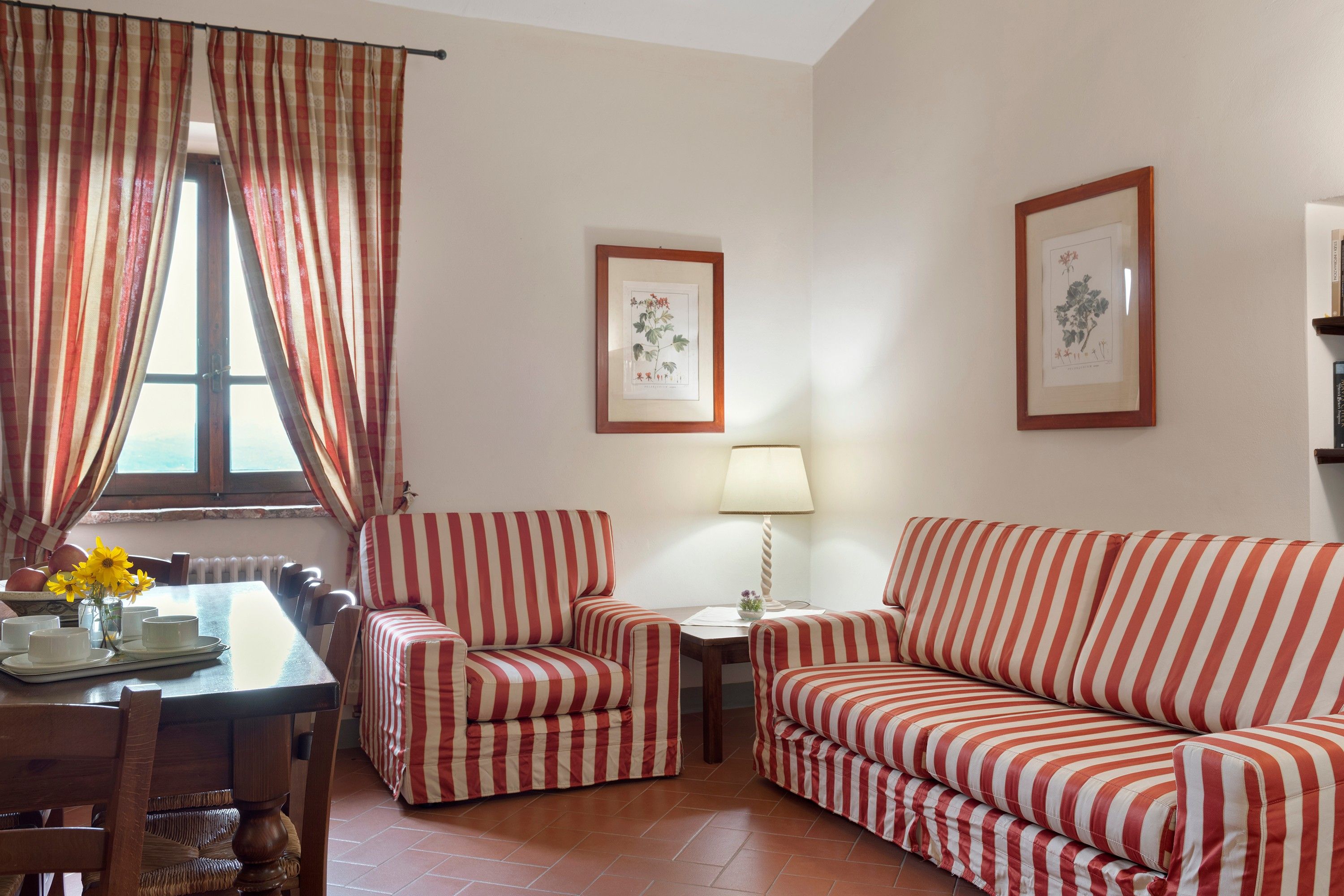 A living room with striped furniture, a dining table with a vase of flowers, and two framed botanical illustrations on the wall.