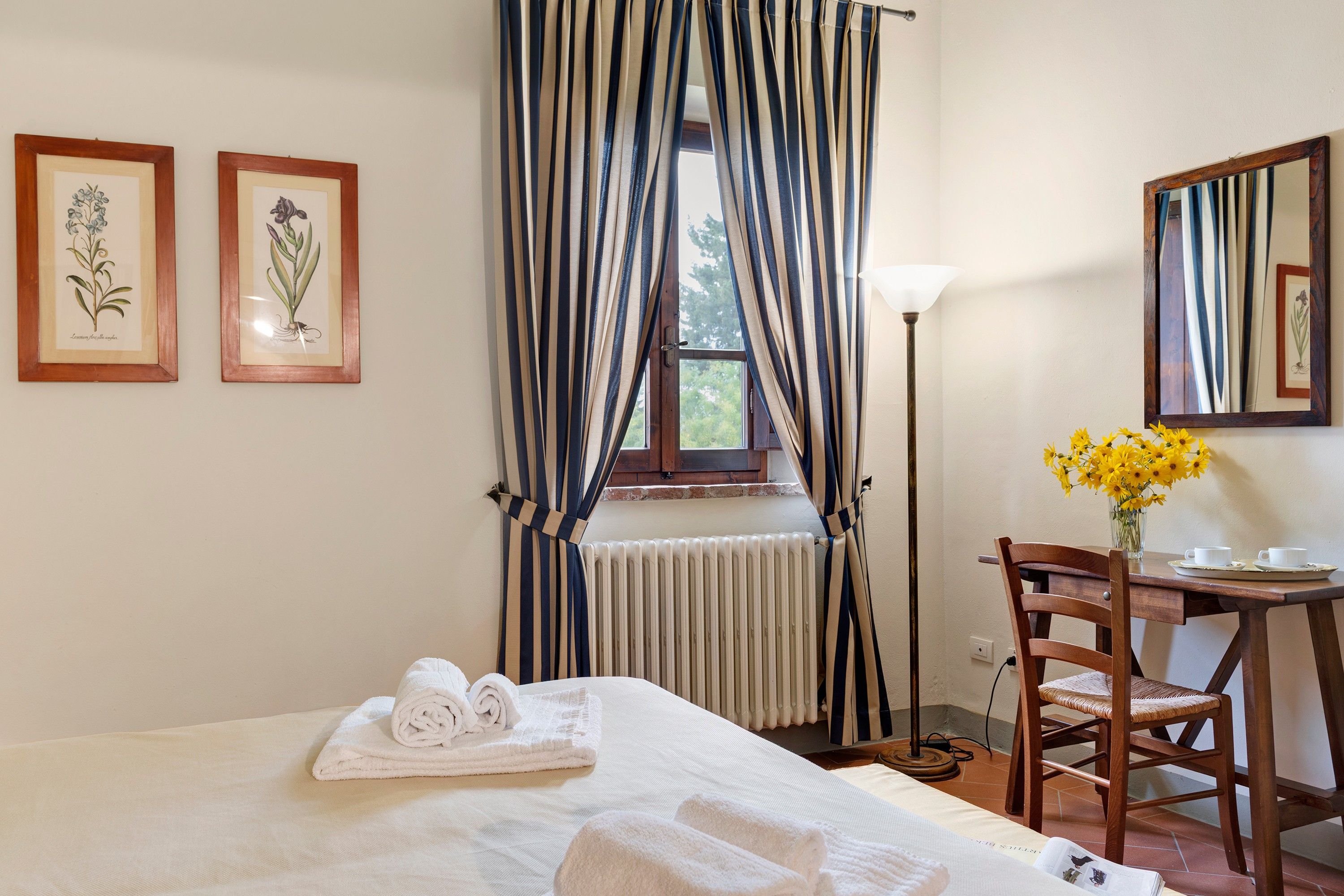 A hotel room with a bed, a chair, a table with a vase of flowers, and two framed botanical illustrations on the wall.
