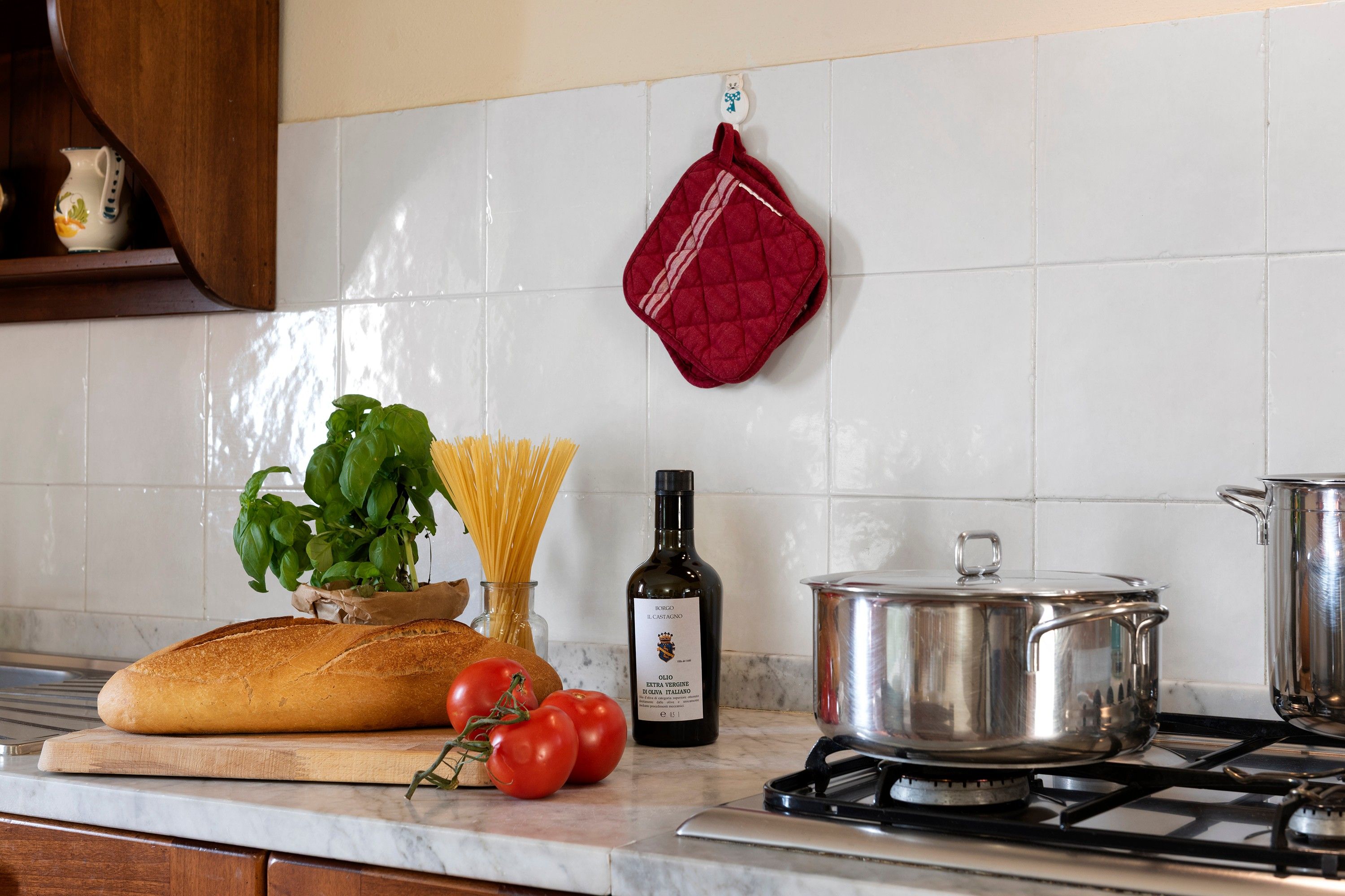 A kitchen counter with a loaf of bread, tomatoes, basil, spaghetti, a bottle of olive oil, and a pot on a stove.