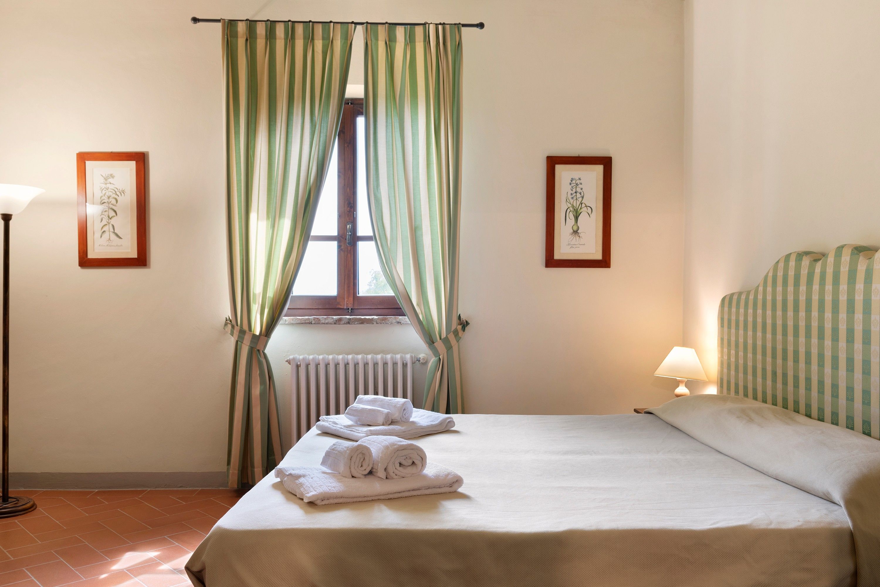 Habitación de hotel con cama, radiador, ventana con cortinas, lámpara y dos ilustraciones botánicas enmarcadas en las paredes.