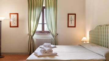 Habitación de hotel con cama, radiador, ventana con cortinas, lámpara y dos ilustraciones botánicas enmarcadas en las paredes.