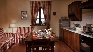 Een eetgedeelte met een houten tafel, stoelen en een rood-wit gestreepte bank. Er is een raam met gordijnen, een keuken met kasten en apparatuur, en een wandplaatje met een plant.