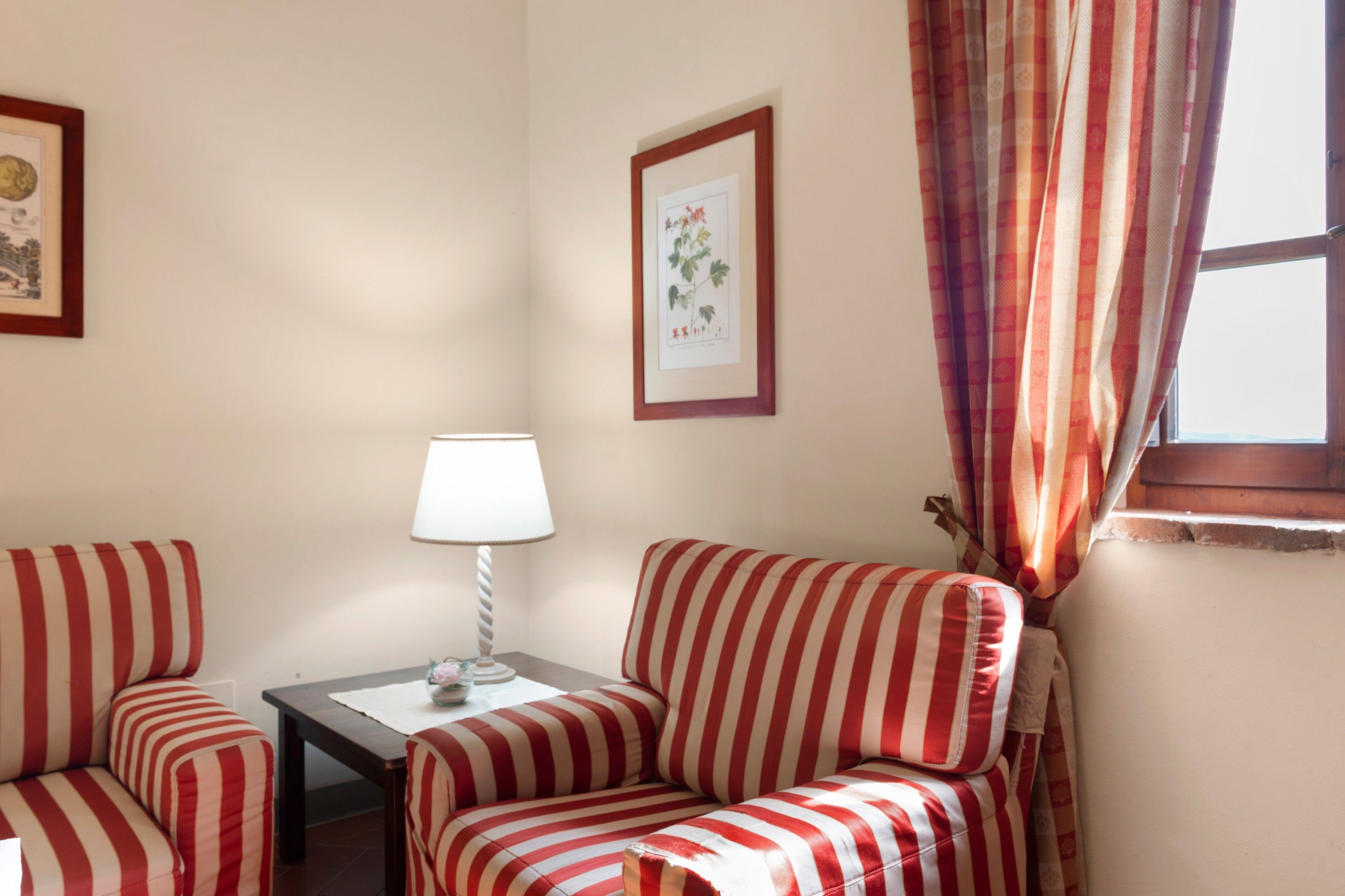 Una habitación con dos sillones a rayas, una mesita con lámpara, una ilustración botánica enmarcada en la pared y una ventana con cortinas.
