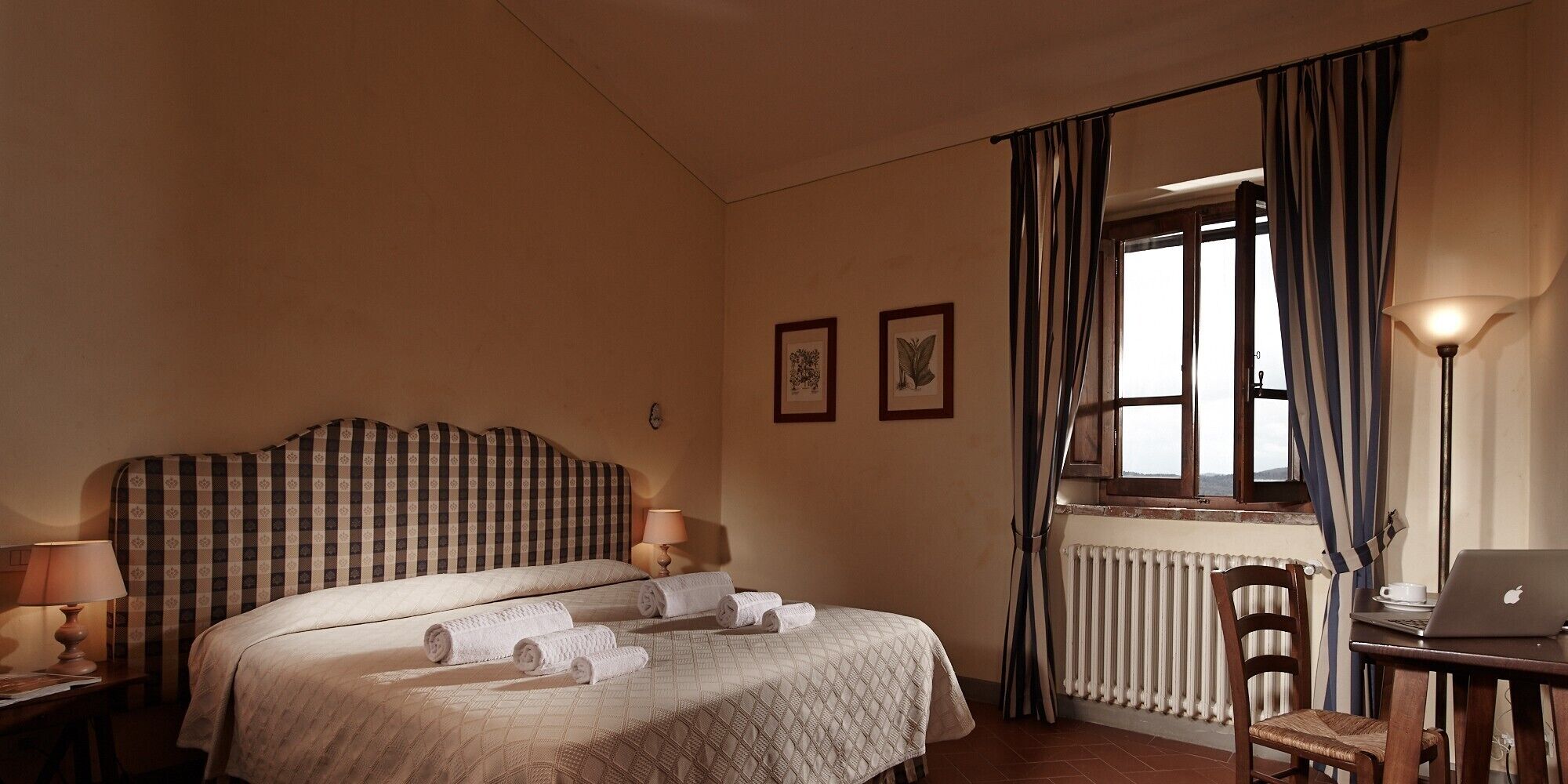 Une chambre d’hôtel avec un lit, un bureau avec un ordinateur portable, un radiateur et une fenêtre avec des rideaux.