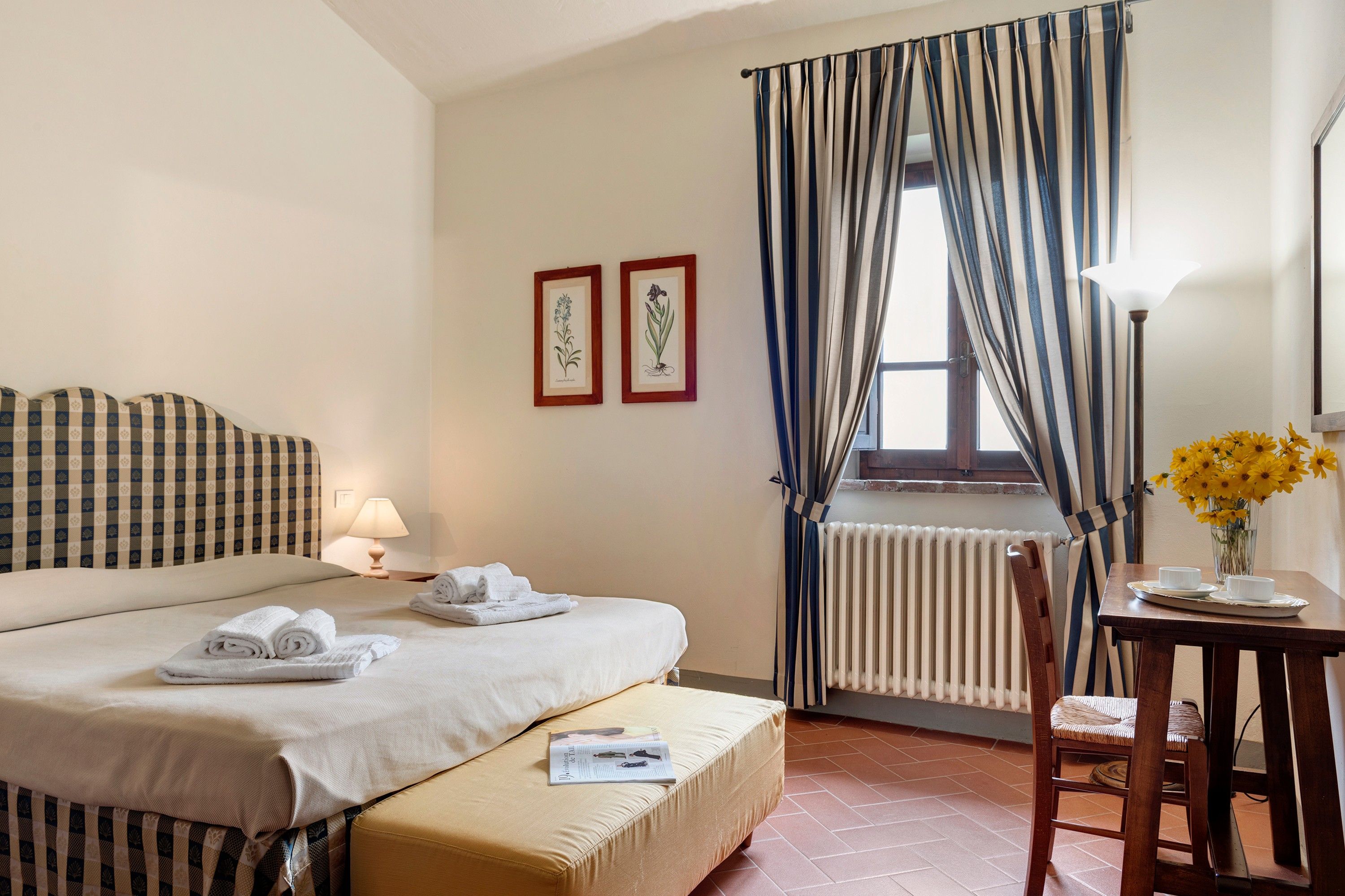 A hotel room with a bed, a bedside table with a lamp, a chair, a small table with a vase of flowers, and a window with curtains.