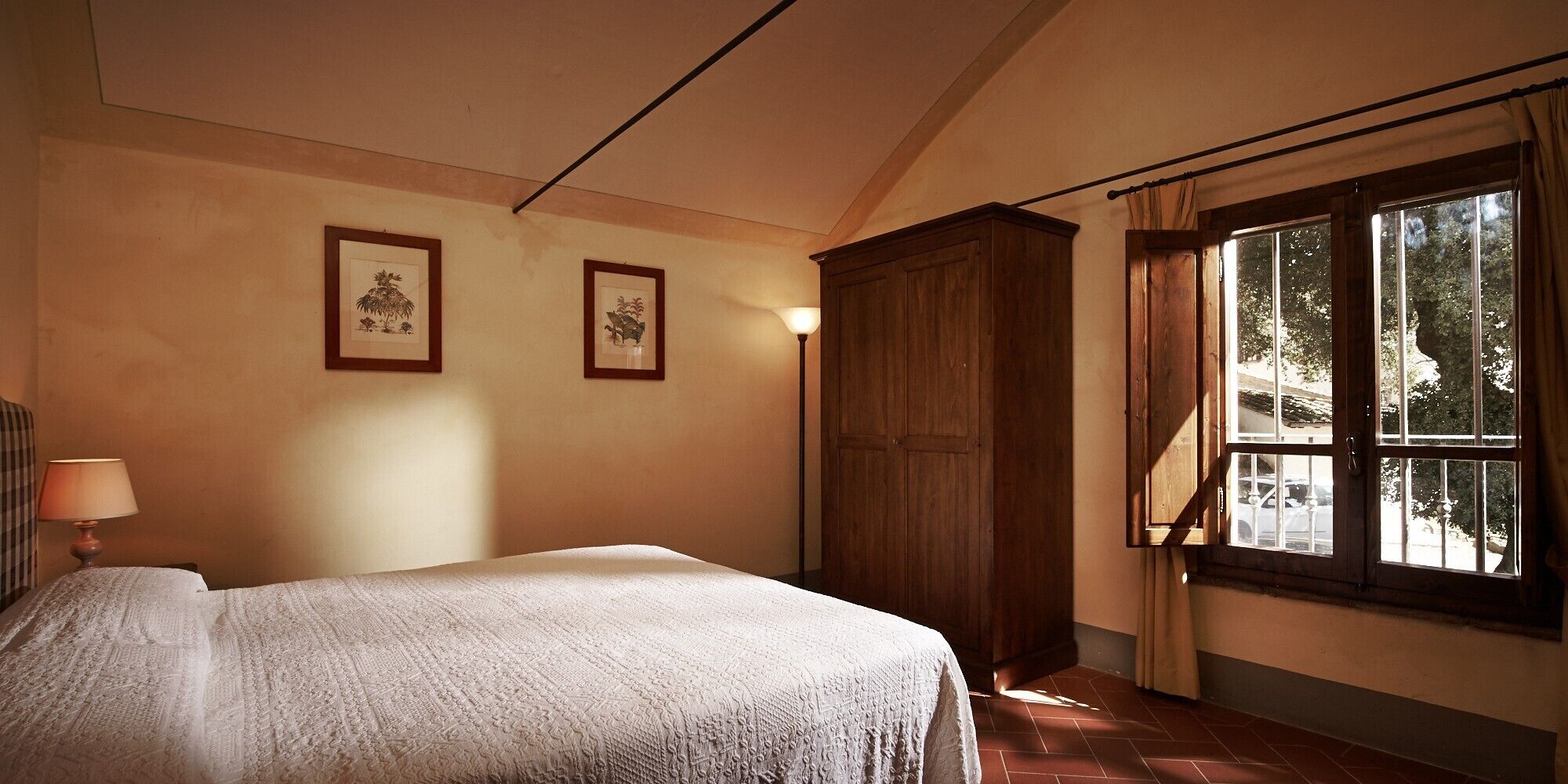 Ein Schlafzimmer mit Bett, Kleiderschrank, Lampe und Fenster mit Blick auf Bäume.