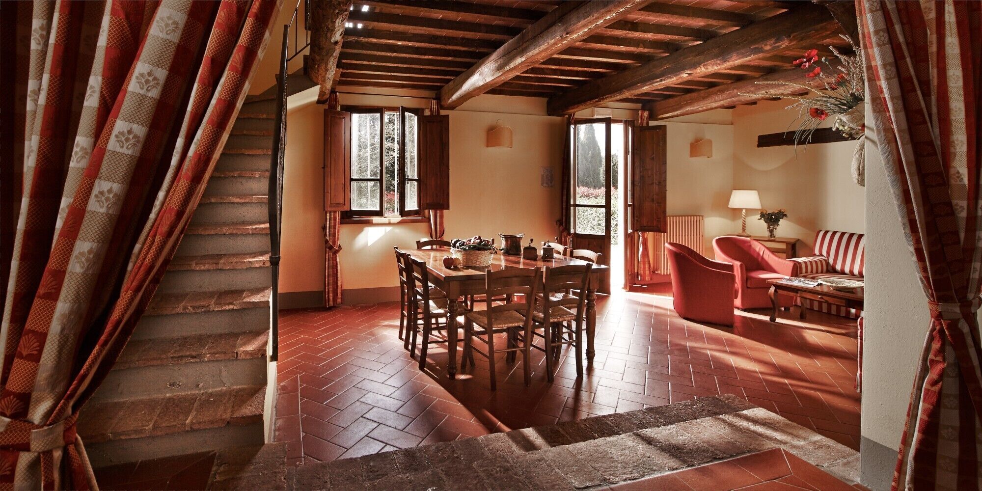 Un interno rustico con una scala in legno, un'area pranzo con tavolo e sedie, e un salotto con divano e poltrone.