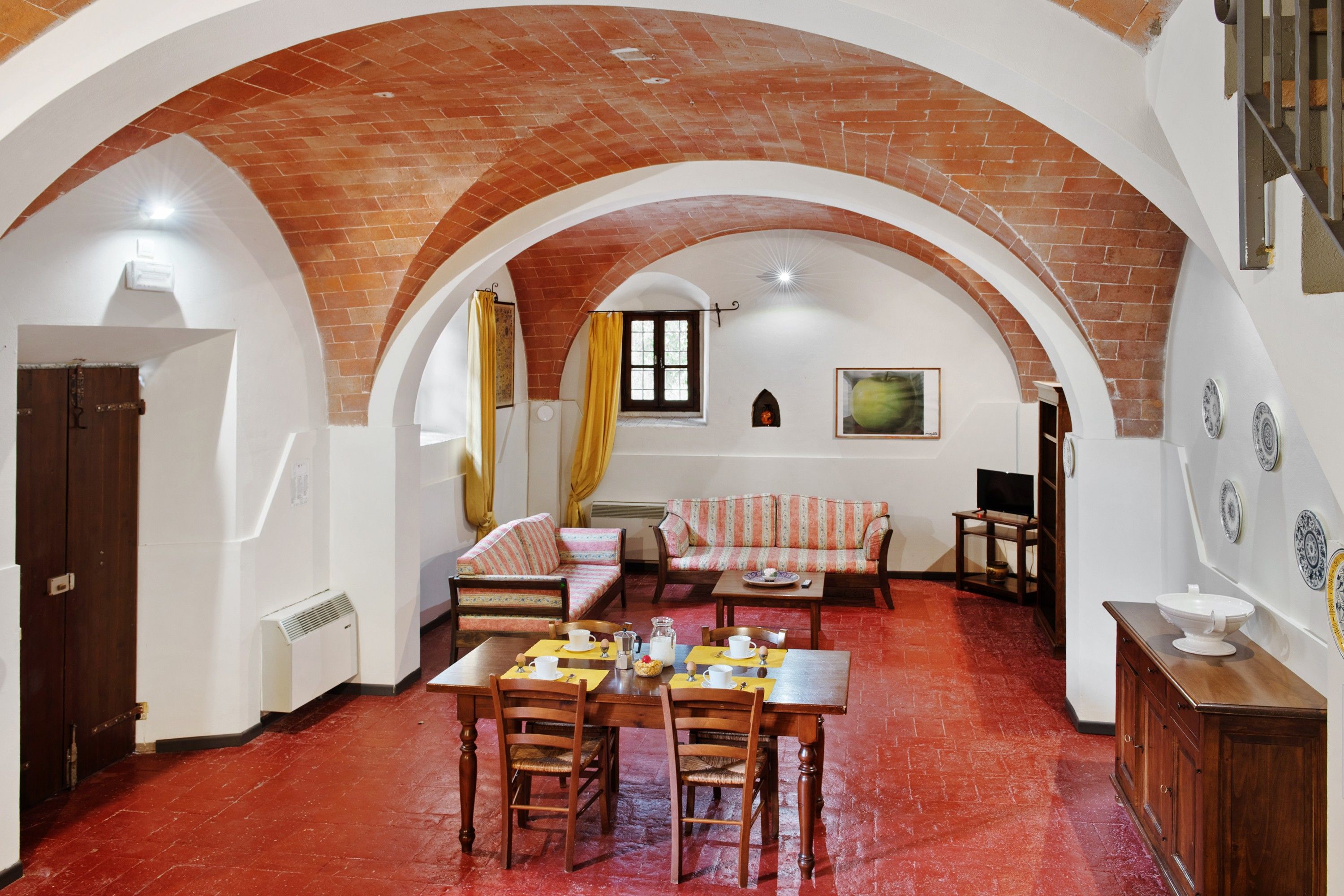 A dining area with a brick floor, a wooden table, and chairs. There is a sofa and a television in the background.