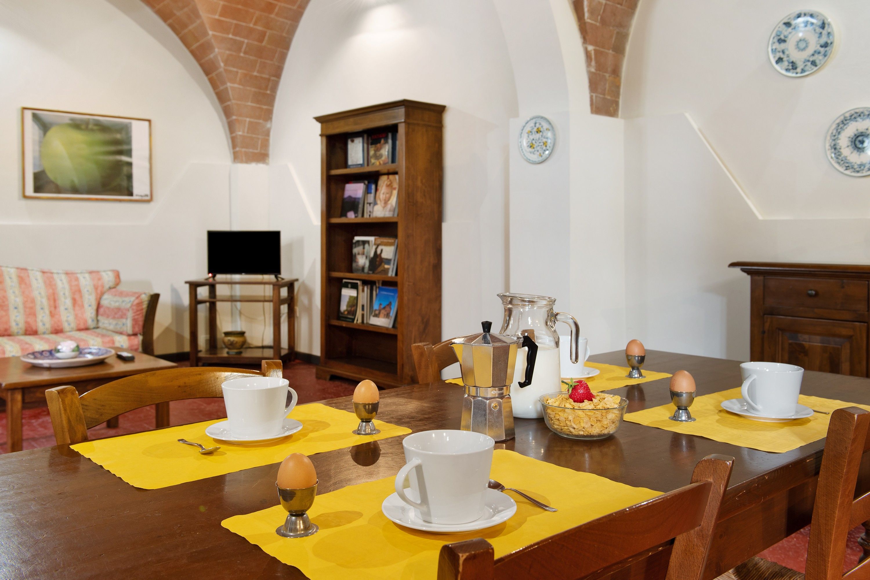 A dining area with a wooden table set for breakfast, a coffee maker, and a bookshelf in the background.