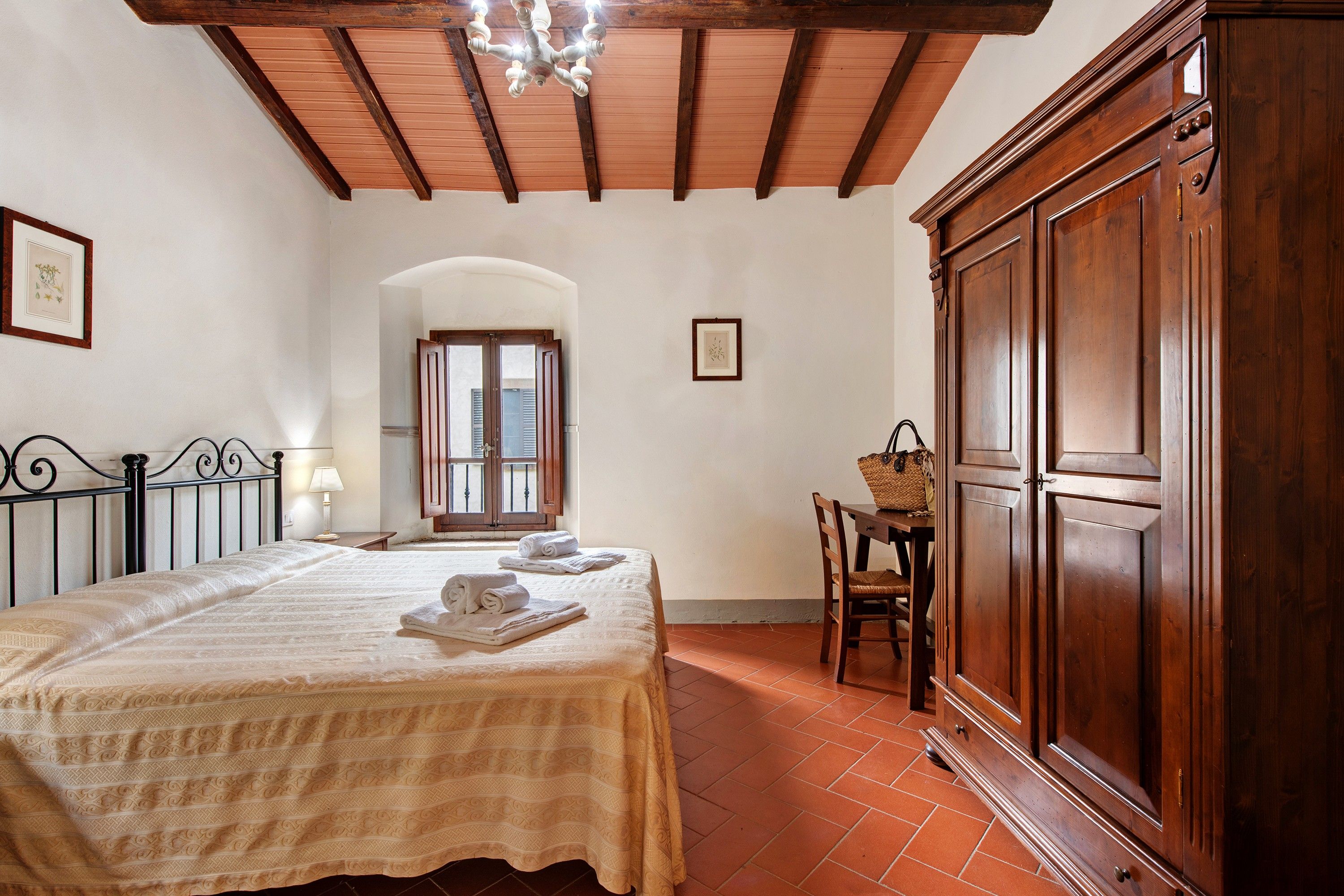 Quarto com cama grande, guarda-roupa de madeira, escrivaninha com cadeira e janela com portadas.