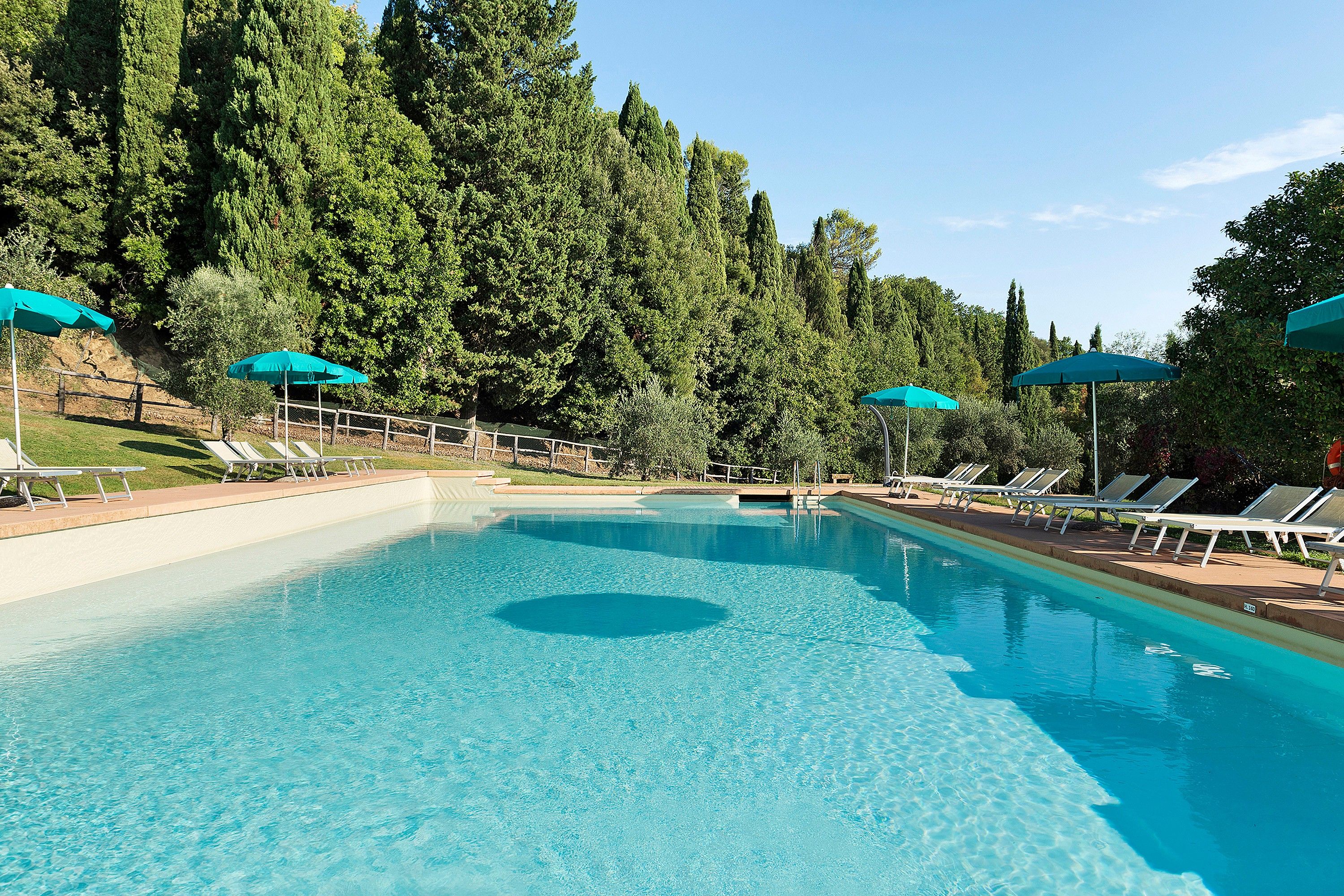 A clear swimming pool with a circular center, surrounded by lounge chairs and umbrellas, set against a backdrop of dense trees.