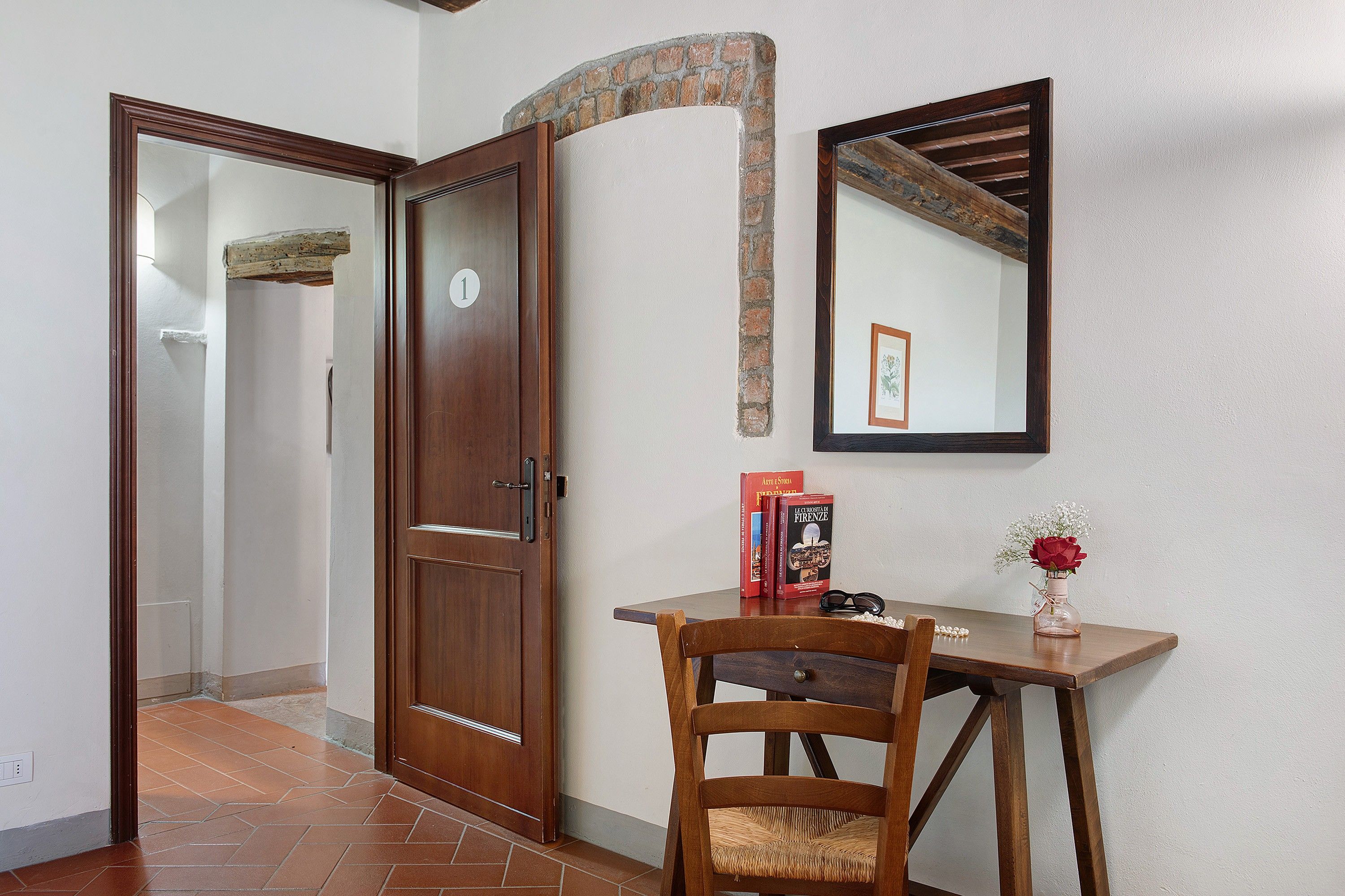 A wooden door with a number '1' sign, a mirror on the wall, and a small table with books and a vase.