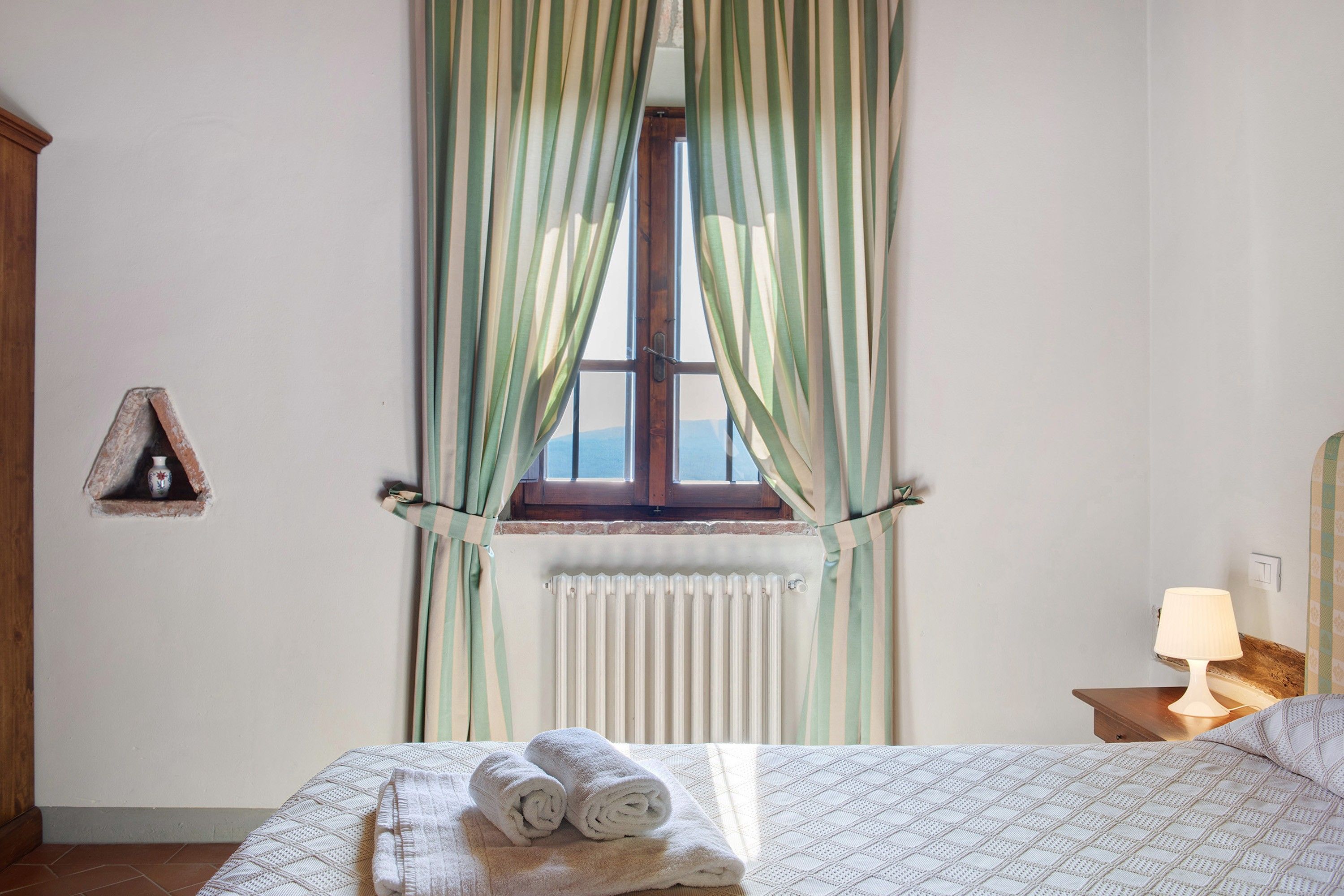 A bedroom with a bed, a window with curtains, a radiator, a bedside table with a lamp, and a small shelf with a decorative item.