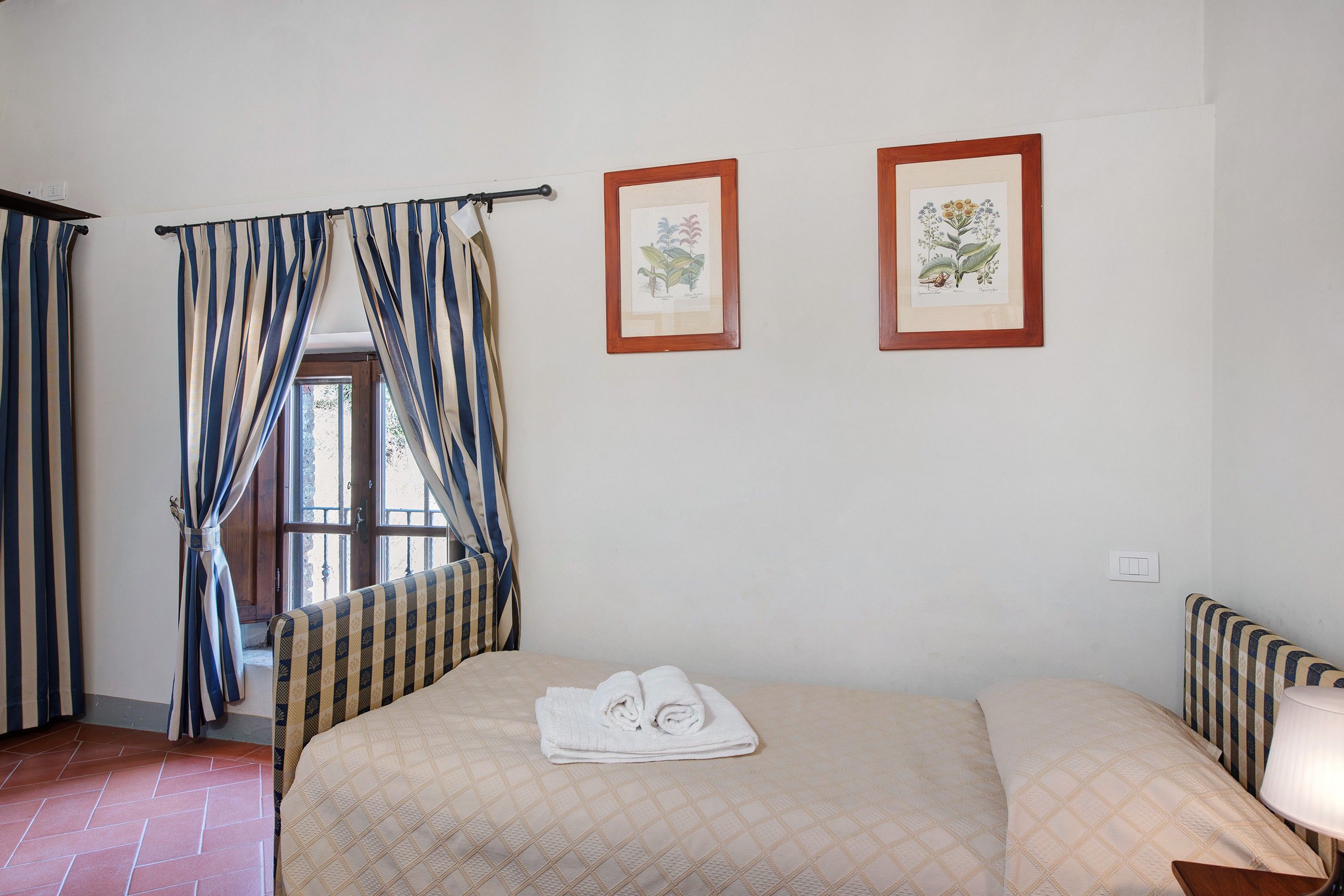 A hotel room with a bed, a window with curtains, and two framed botanical illustrations on the wall.
