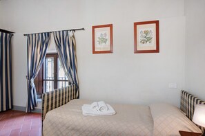 Una habitación de hotel con una cama, un ventanal con cortinas y dos ilustraciones botánicas enmarcadas en la pared.