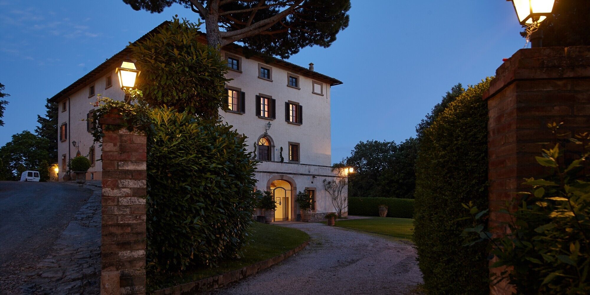 A two-story building with a paved driveway leading to a gated entrance, surrounded by well-maintained hedges and a clear sky.