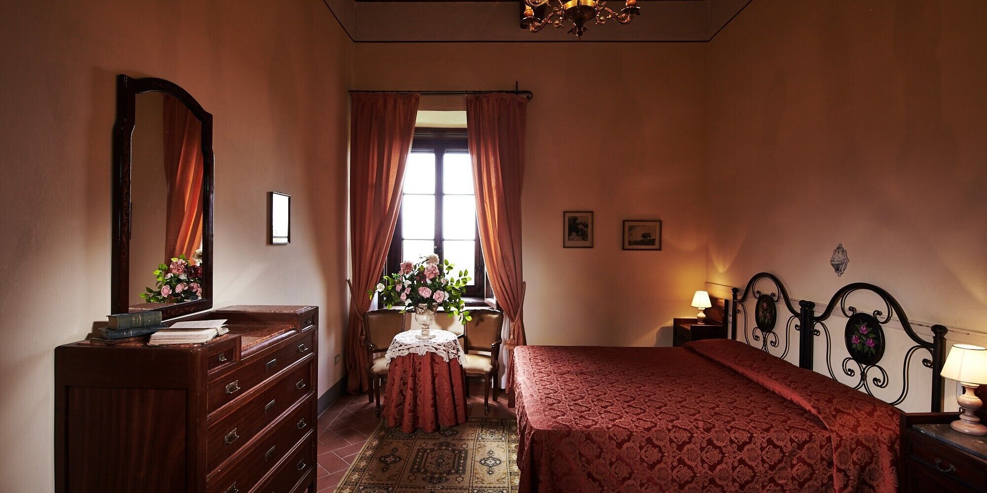 Een slaapkamer met een bed, een dressoir, een bureau met stoel, een raam met gordijnen en een kroonluchter.