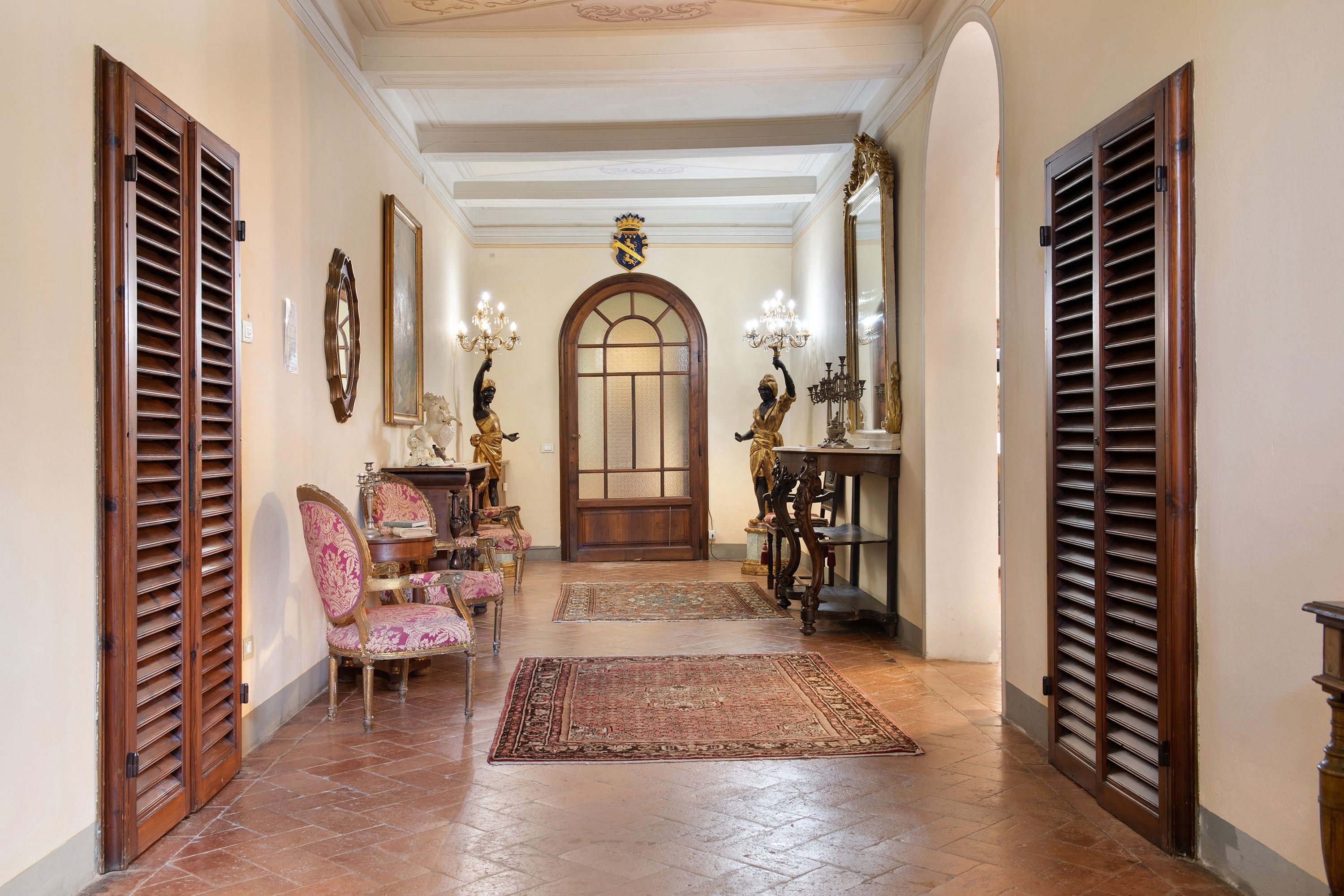 A hallway with a grand arched doorway, flanked by wooden shutters, leading to a room with a large mirror and a statue.