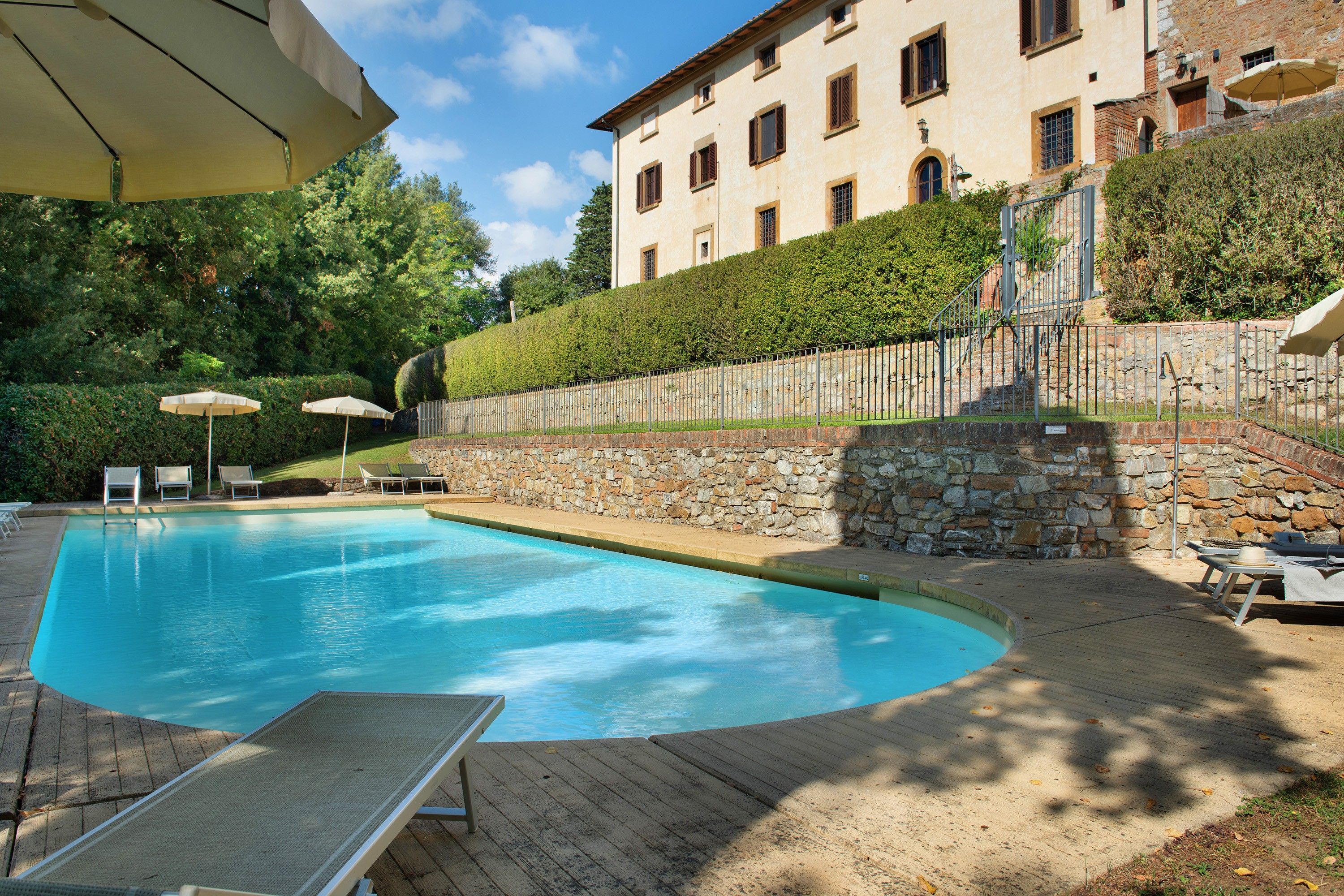 A swimming pool with lounge chairs and umbrellas, surrounded by a stone wall and a building with multiple windows.