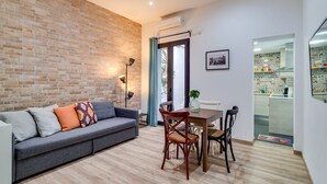 A living room with a grey sofa, a dining table with chairs, a brick wall, and a kitchen visible through an open door. - GuestReady - Charming Studio with Patio in Gràcia (Barcelona)