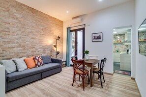 Ein Wohnzimmer mit grauem Sofa, einem Esstisch mit Stühlen, einer Ziegelwand und einer durchgehenden Tür erkennbaren Küche.