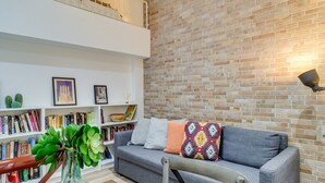A modern living room with a grey sofa, a bookshelf, and a brick wall. - GuestReady - Charming Studio with Patio in Gràcia (Barcelona)