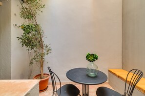 Un petit espace extérieur aménagé avec une table ronde, deux chaises, un pot de fleurs et un banc.