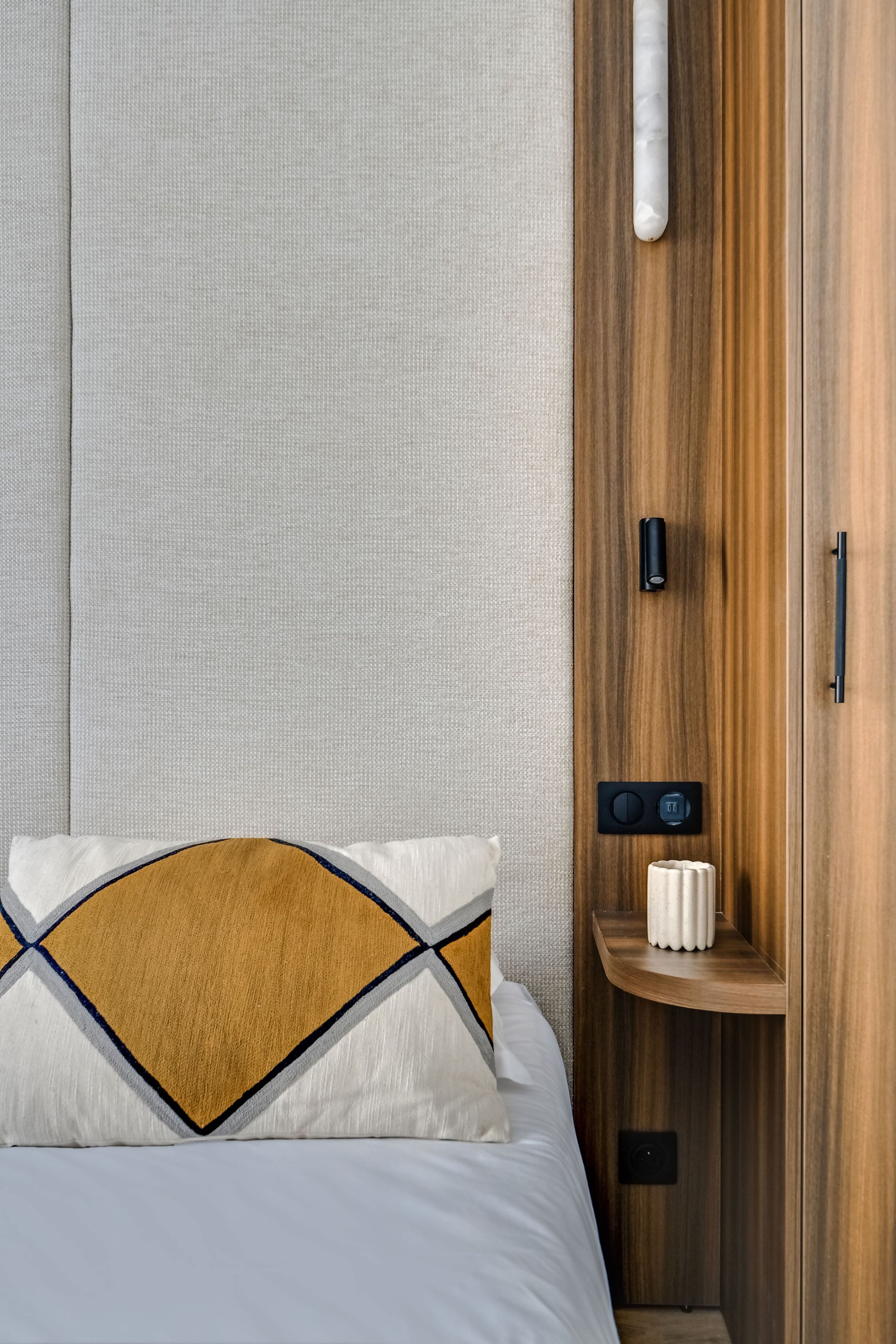 A bedroom with a bed, a wooden wardrobe, a bedside table with a lamp, and a wall-mounted control panel.