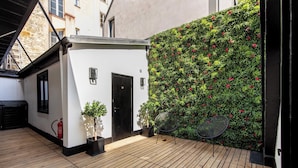 Un patio con cubierta de madera, un edificio blanco con puerta negra y una pared verde con flores rojas.