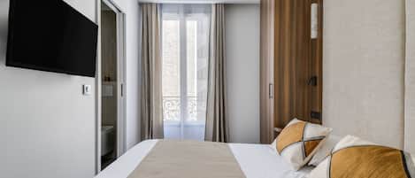 A hotel room with a bed, a TV mounted on the wall, a wardrobe, and a view of a building through the window.