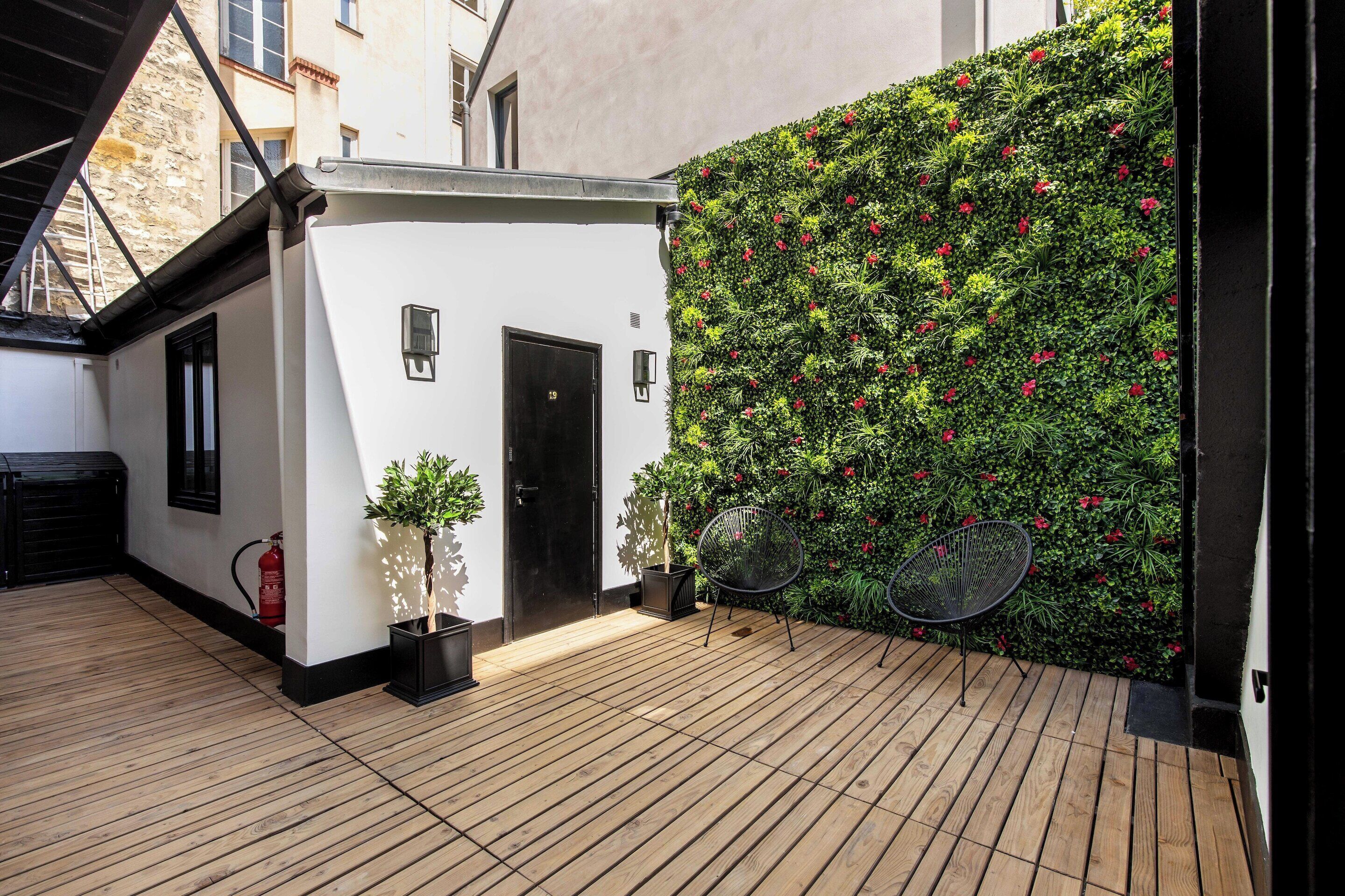 Eine Holzterrasse mit einem weißen Gebäude, einer schwarzen Tür und einer Wand, die mit grünem Blätterwerk und roten Blüten bedeckt ist.