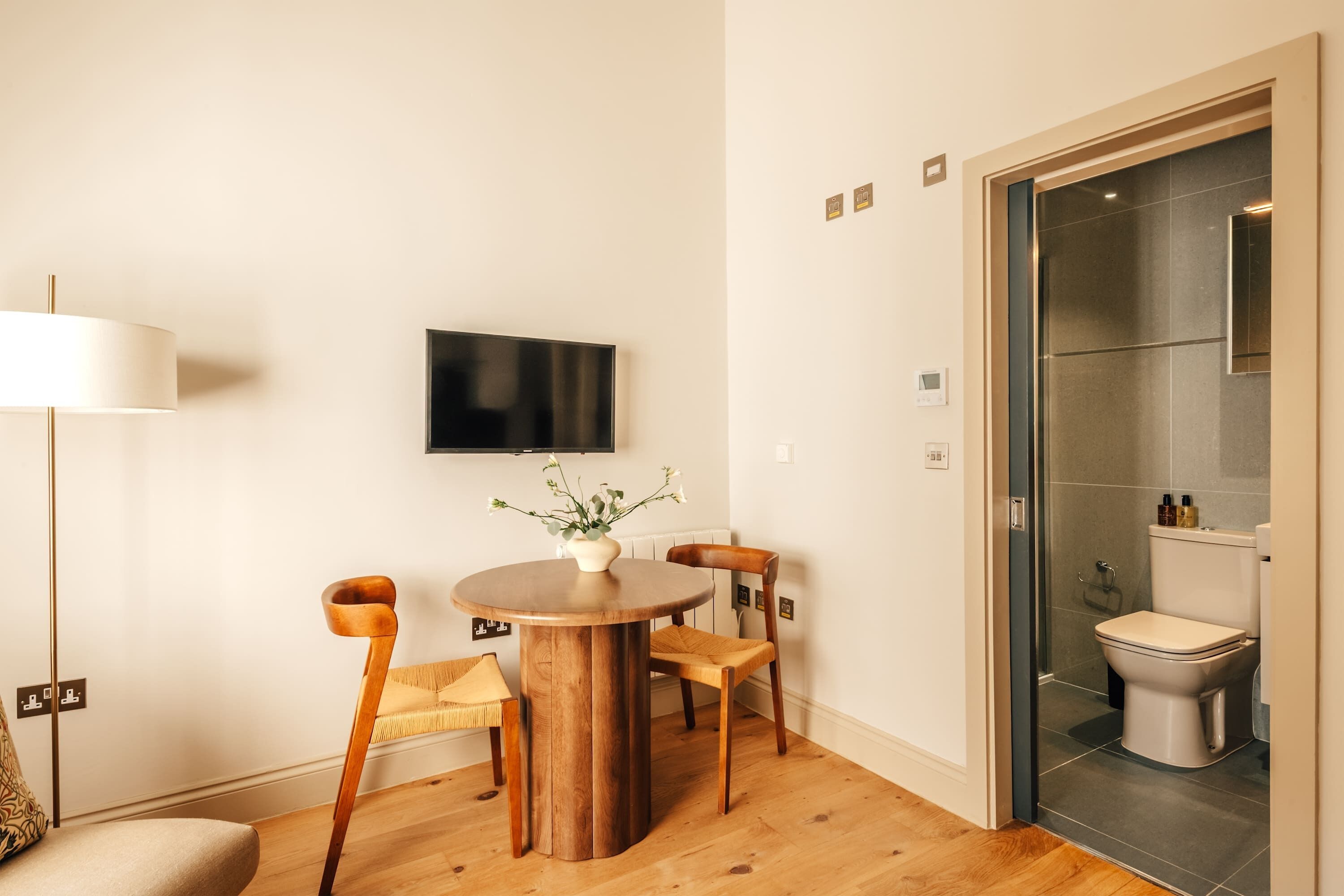 A modern living room with a round wooden table, two chairs, a floor lamp, and a television mounted on the wall.