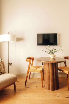 Ein modernes Wohnzimmer mit einem runden Holztisch, zwei Holzstühlen, einem Sofa, einer Stehlampe und einem an der Wand befestigten Flachbildfernseher.