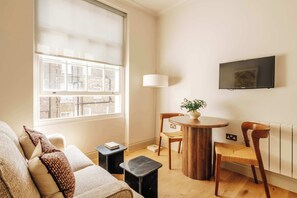 A cozy living room with a large window, a round wooden table, a sofa, and a TV mounted on the wall. - The Cheniston | 1-Bed Apartment Kensington (London)