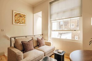 A modern living room with a sofa, a small table, and a large window with a view of a brick building. - The Cheniston by nestor | Apartment Kensington (London)