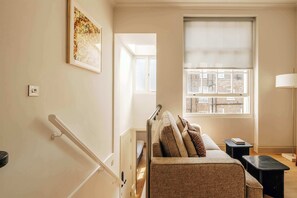 A bedroom with a bed, a window with a view of buildings, a door, and a light switch. - The Cheniston | 1-Bed Apartment Kensington (London)