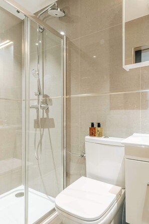 Une salle de bain moderne avec une douche à parois de verre, des toilettes blanches et une petite étagère contenant des articles de toilette.