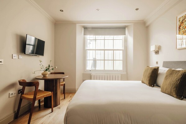 A hotel room with a bed, a desk with a chair, a television, and a window with a view. - The Cheniston | Kensington Lower Ground Studio (London)