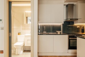 A modern kitchen with white cabinets, a black oven, and a stainless steel range hood. - The Cheniston | Kensington Lower Ground Studio (London)