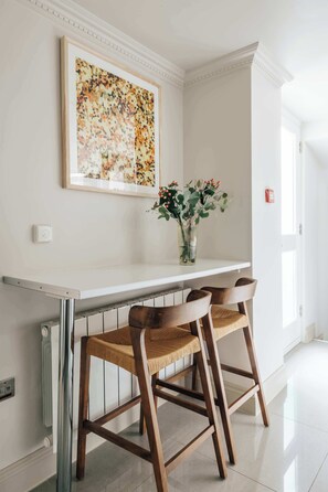 Ein Essbereich mit einem weißen Tisch, zwei Holzsesseln, einer Blumenvase und einem gerahmten Kunstwerk an der Wand.