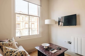 Ein Zimmer mit großem Fenster, einer Couch, einem Tisch mit Fernbedienung und einem an der Wand befestigten Fernseher.