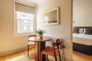 Ein Zimmer mit einem runden Holztisch, zwei Stühlen, einer Küstpflanze, einem Bett mit Kissen und einem Fenster mit Blick auf Gebäude.