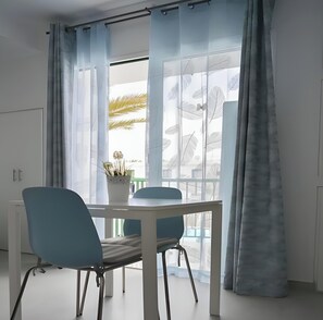 Una habitación moderna con una mesa de comedor y sillas, una planta en maceta y un ventanal con cortinas translúcidas.