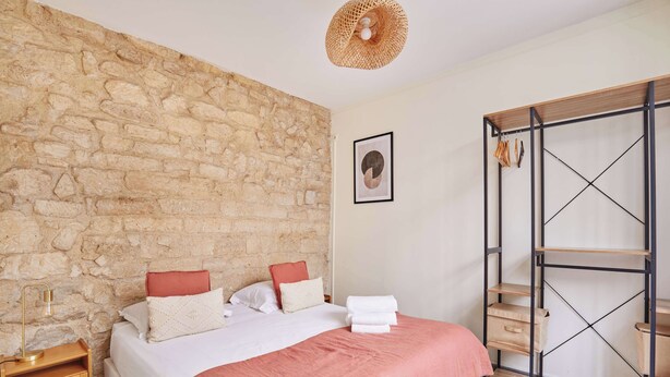 Ein Schlafzimmer mit einer Steinswand, einem Bett mit rosa Decke, einem Holz-Nachttisch, einem Metallschrank und einer Pendelleuchte.