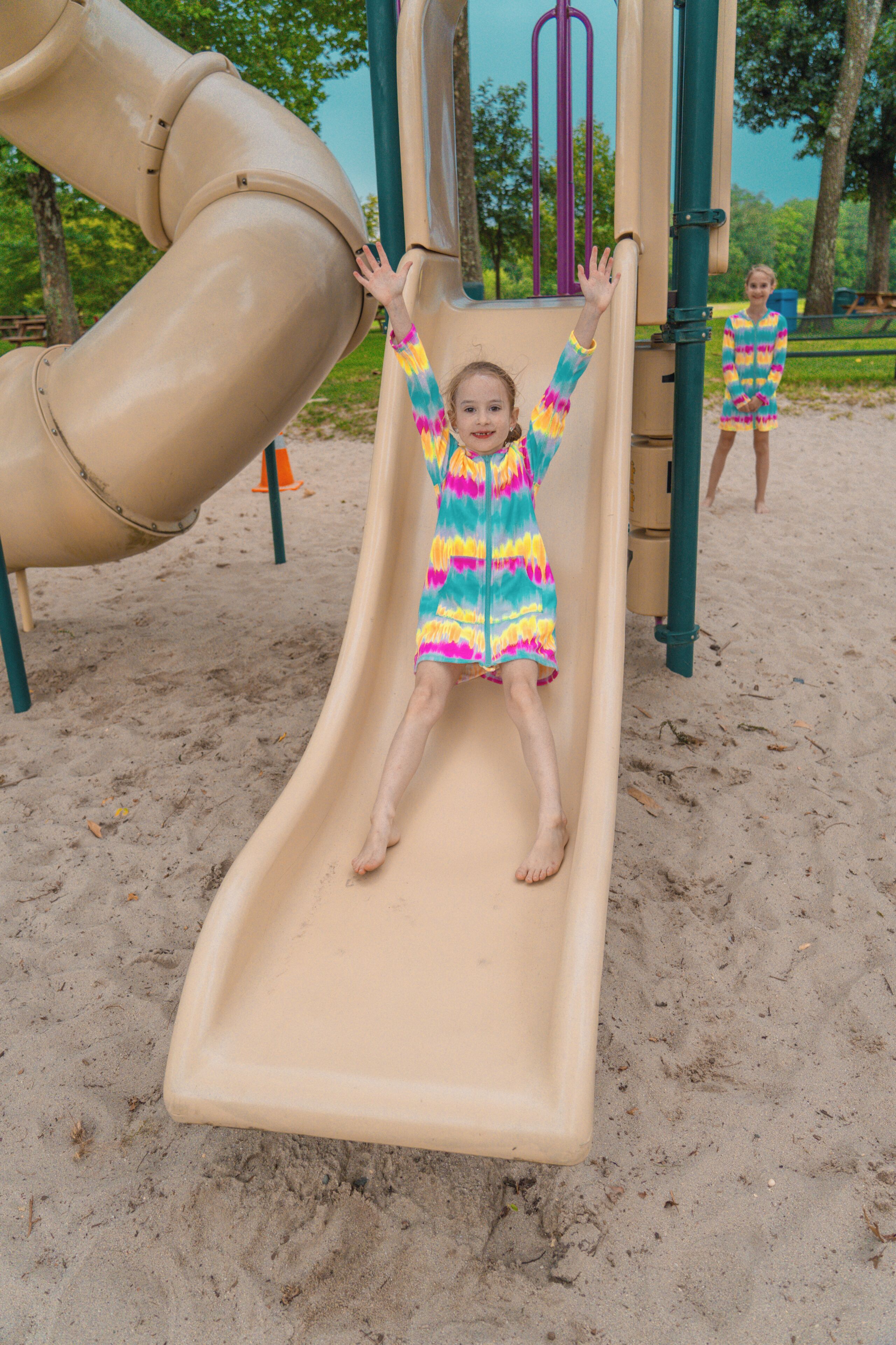 A child sliding down a beige slide at a playground with a sandy ground and trees in the background.